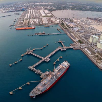 Aerial view of an LNG tanker docked at an offshore terminal with multiple pipelines, storage tanks, and an industrial complex. Captures global energy logistics